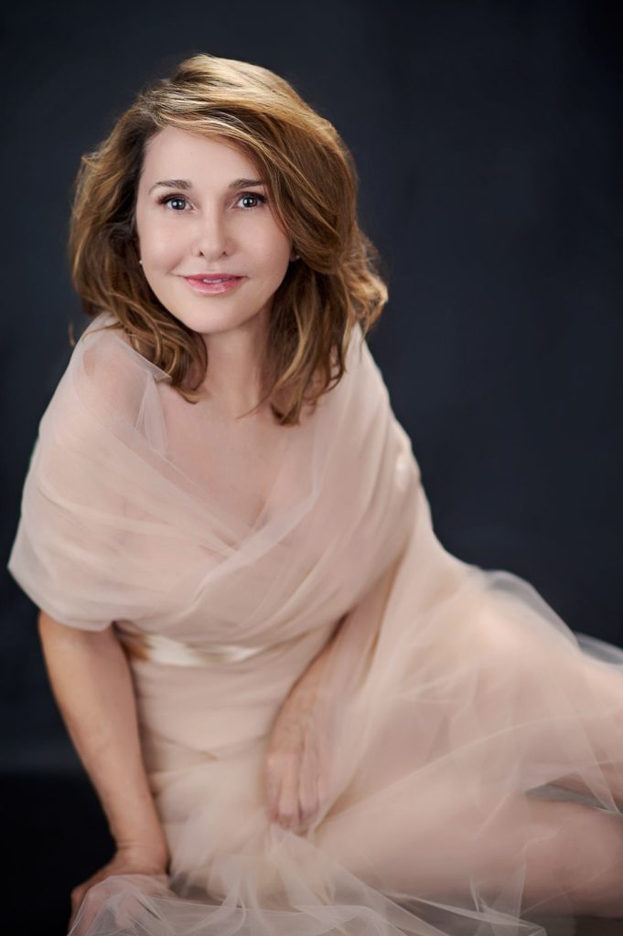 Soft boudoir-style portrait of woman wrapped in blush tulle on a dark background.