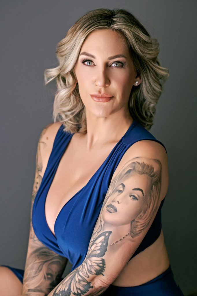 Close-up studio portrait of woman in blue dress with tattoos on a neutral background.