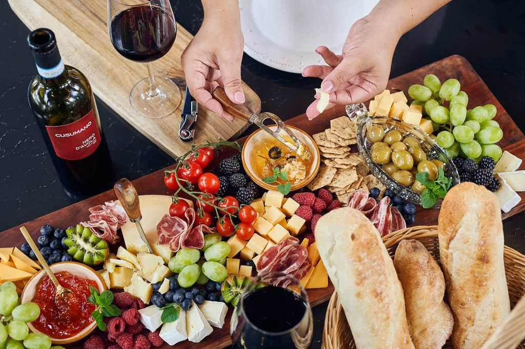 Overhead food photo of a charcuterie board with cheese, fruit, bread, and wine