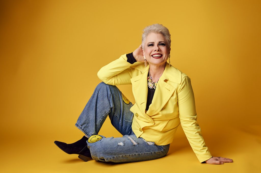 Studio portrait of woman in yellow jacket and jeans sitting on a bright yellow background.