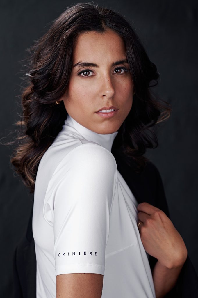Moody studio headshot of brunette woman in white top on a dark background.