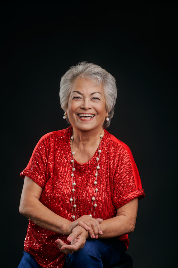 Joyful studio portrait of older woman in red sequins smiling against a dark background.