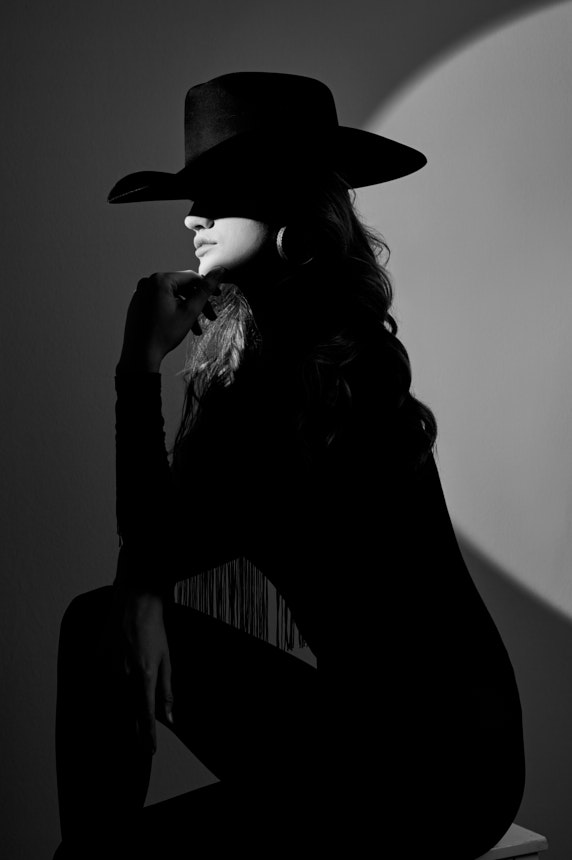 Black-and-white editorial portrait of woman in wide-brim hat with dramatic side lighting.