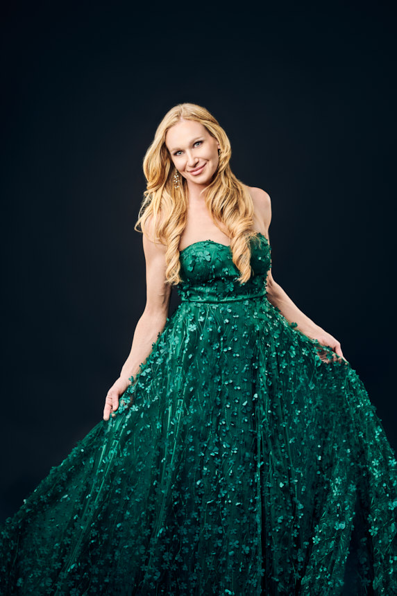 Studio portrait of woman in emerald green gown holding her skirt on a dark background.